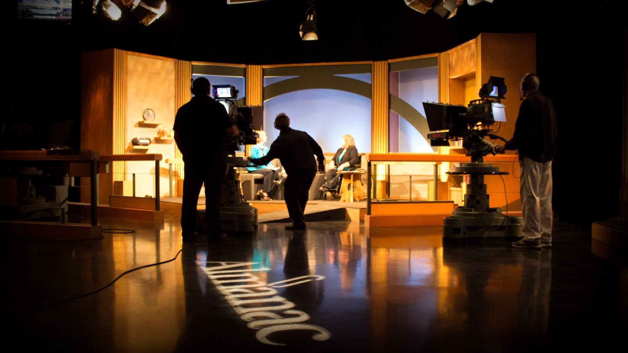 Television studio set with two hosts seated and three crew members operating cameras and equipment, with the word "Almanac" projected on the shiny floor.